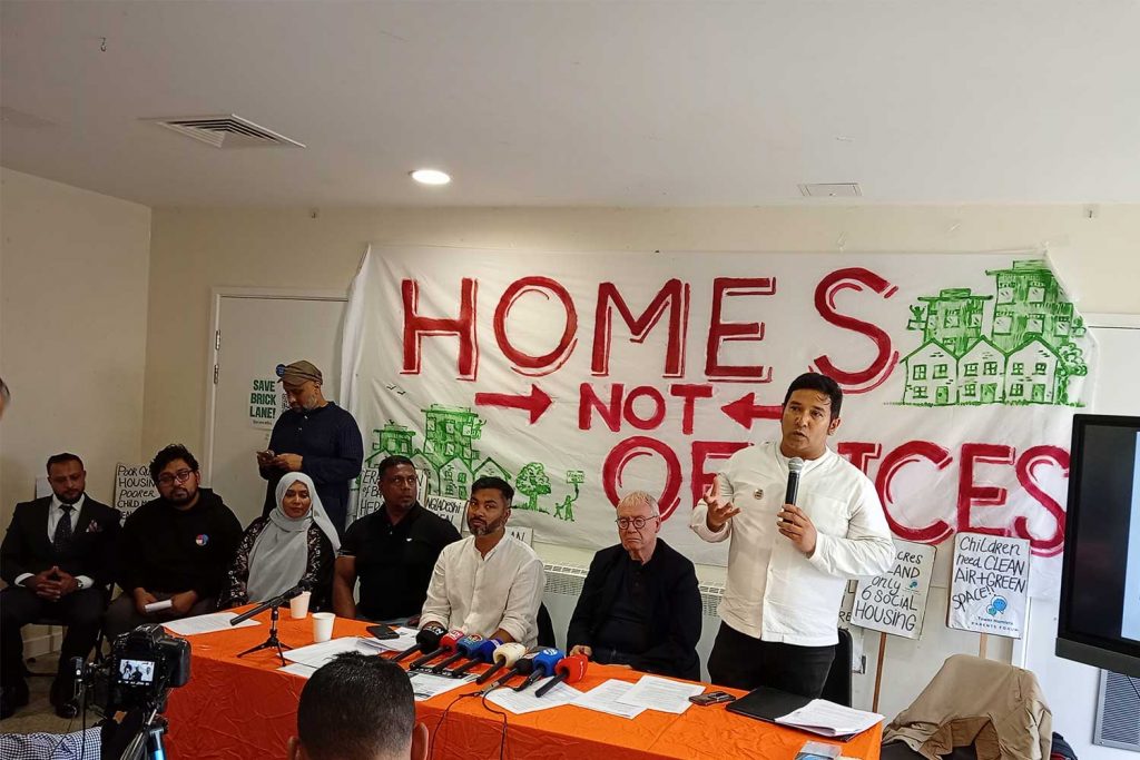 A group of people sit in front of a 'homes not offices' banner with microphones. One speaker stands up and speaks into a microphone, Brick Lane, east London