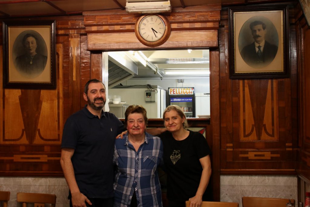Nev, Maria and Anna smiling together, with the portraits of their grandparents - the founders of E Pellicci - either side of them, inside the cafe in Bethnal Green