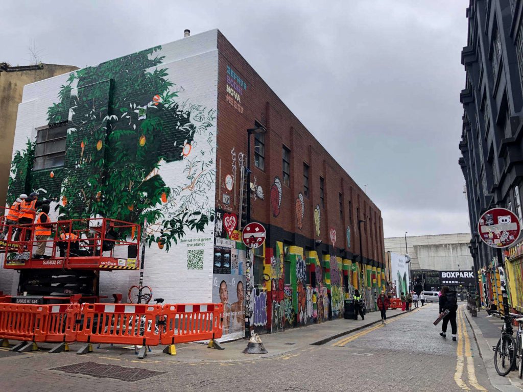 A commercial mural is being painted onto the side of a wall of a Zapp warehouse to advertise Treedom. In the background there is the food and drinks containers venue, Boxpark.