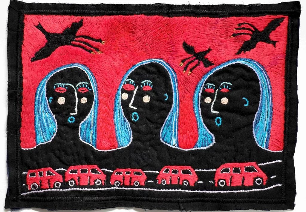 Piece of embroidered material with three abstract faces above a road with five red vehicles and birds fly above their heads.