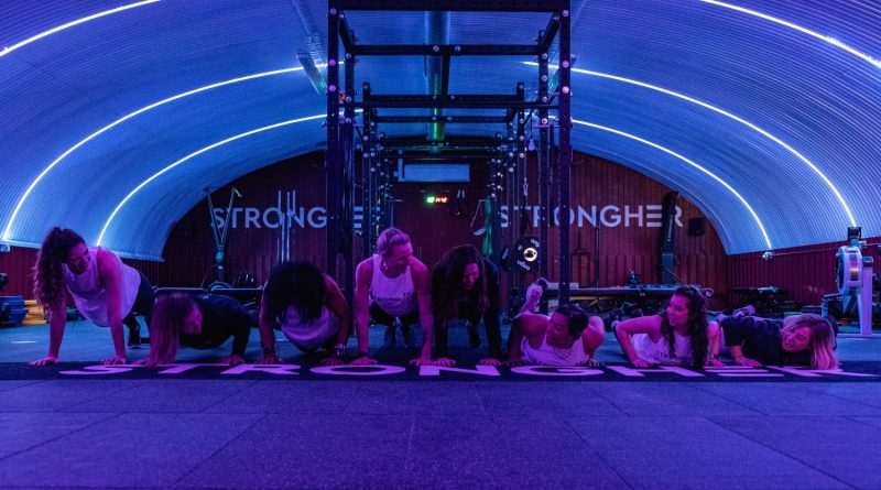 A row of women are in plank position with the background of StrongHer strength and conditioning space